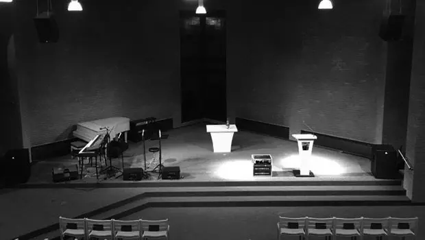 Schwarzweißbild eines Auditoriums in einer Kirche mit weißem Klavier, Keyboard, Lautsprechern, weißem Rednerpult und kleinem PA-Equipment.