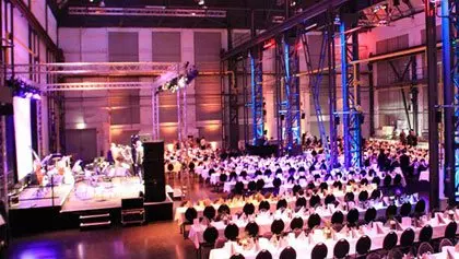 Umgebaute Fabrikhalle als sehr großer Veranstaltungsraum mit Bühne und Bestuhlung im rose´rotem Licht.