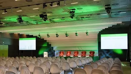 Sehr großer Veranstaltungsraum mit Bühne, weißen Schalensitzen als Bestuhlung, Lichttraversen und 2 sehr großen Videowänden im grünen Licht.
